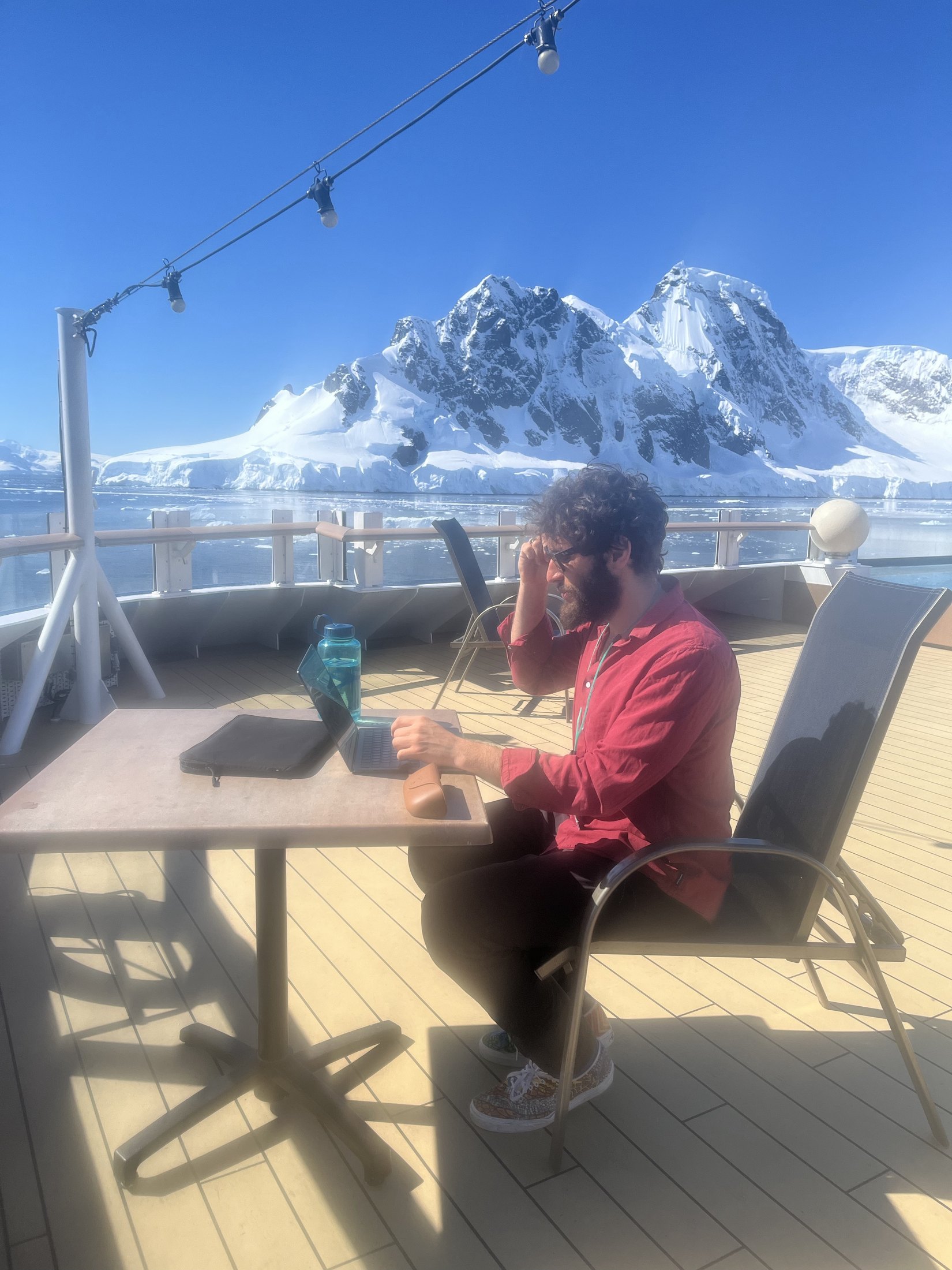 Mathew Youssef in a red shirt working at a laptop on a ship's deck, snow-covered Antarctic peninsula mountains across the water behind him.