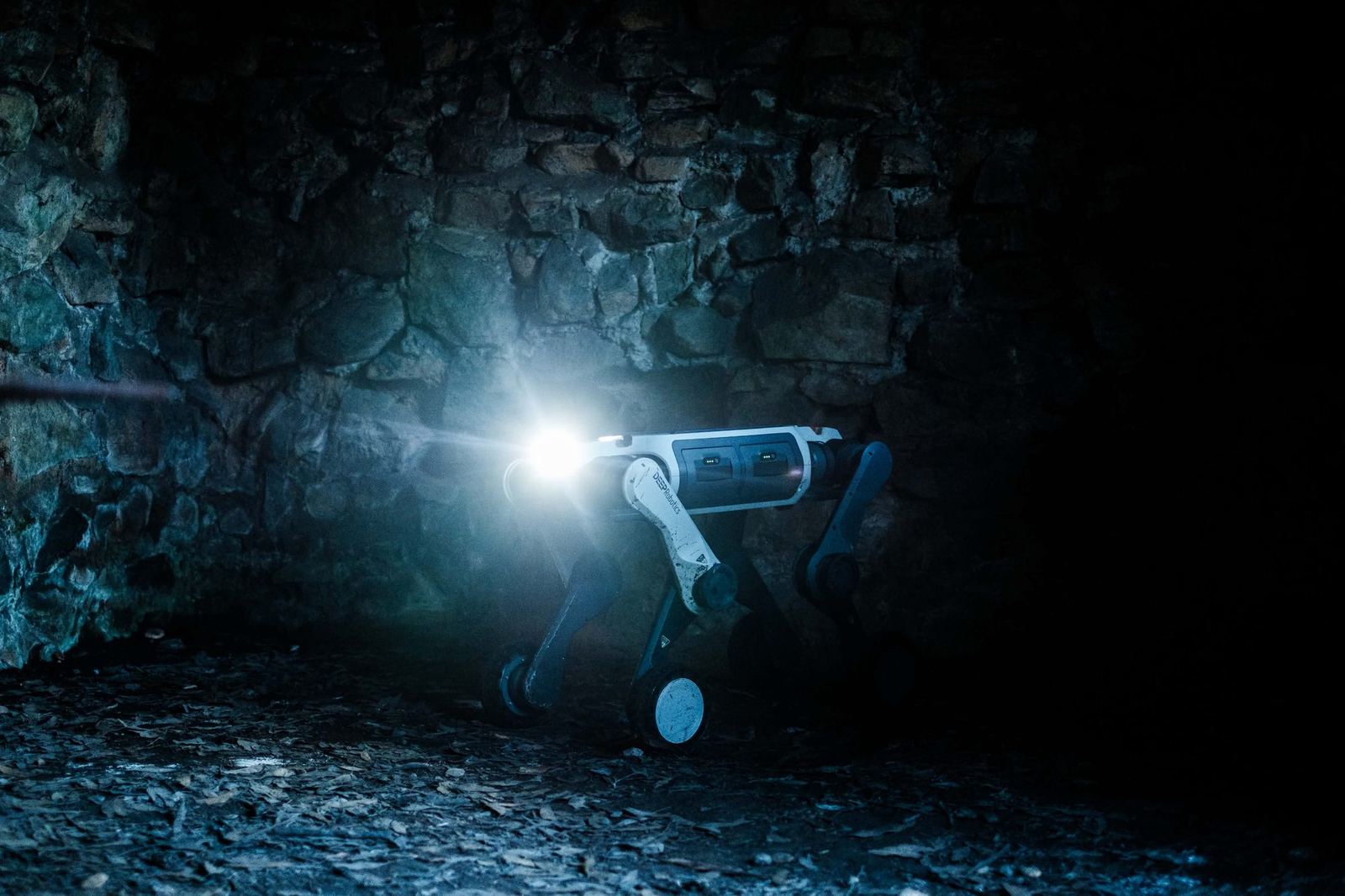 A wheeled-leg quadruped stands in a dark stone interior, its headlight throwing a beam across the rock wall.