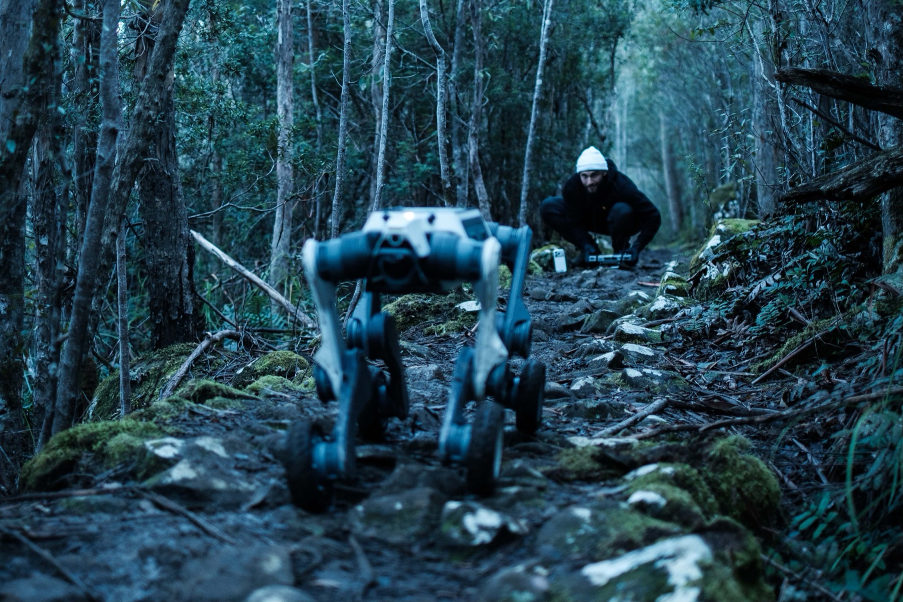 Blue hour on a rocky forest trail: the wheeled-leg ground platform walks toward the camera while Mathew Youssef crouches on the track behind it, controller in hand.