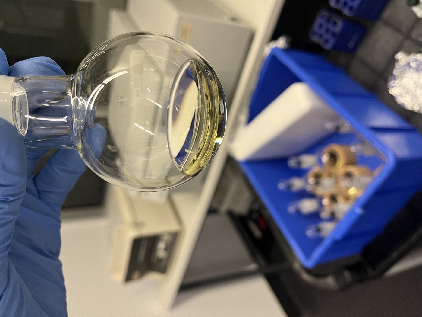 A gloved hand holds a round-bottom flask of pale gold extract above a bin of sample vials.