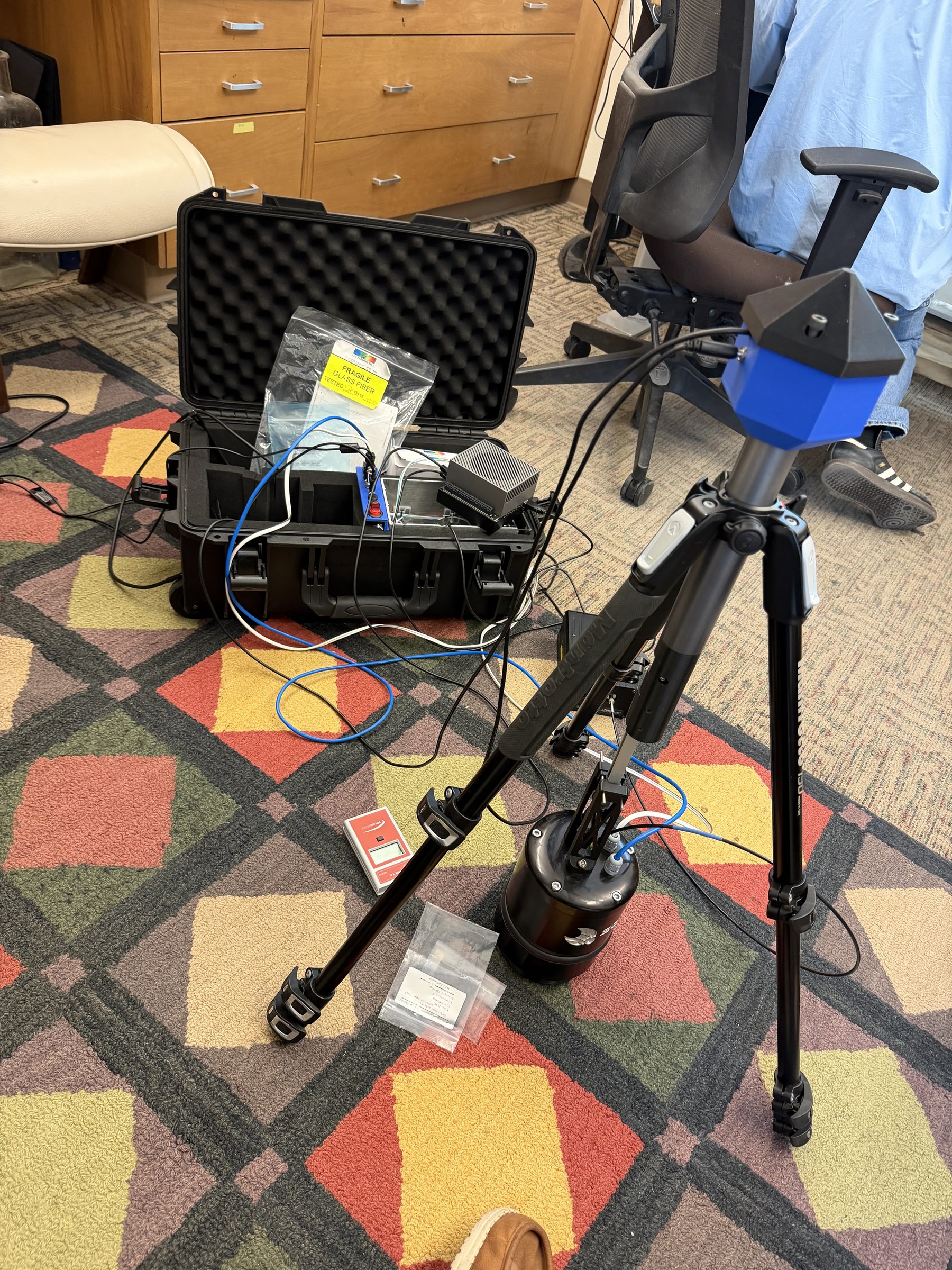 The Spectrophage prototype set up on an office floor: a tripod-mounted sensor head above a black light-trapped dome unit, fibre optics running to a foam-lined transport case of electronics.