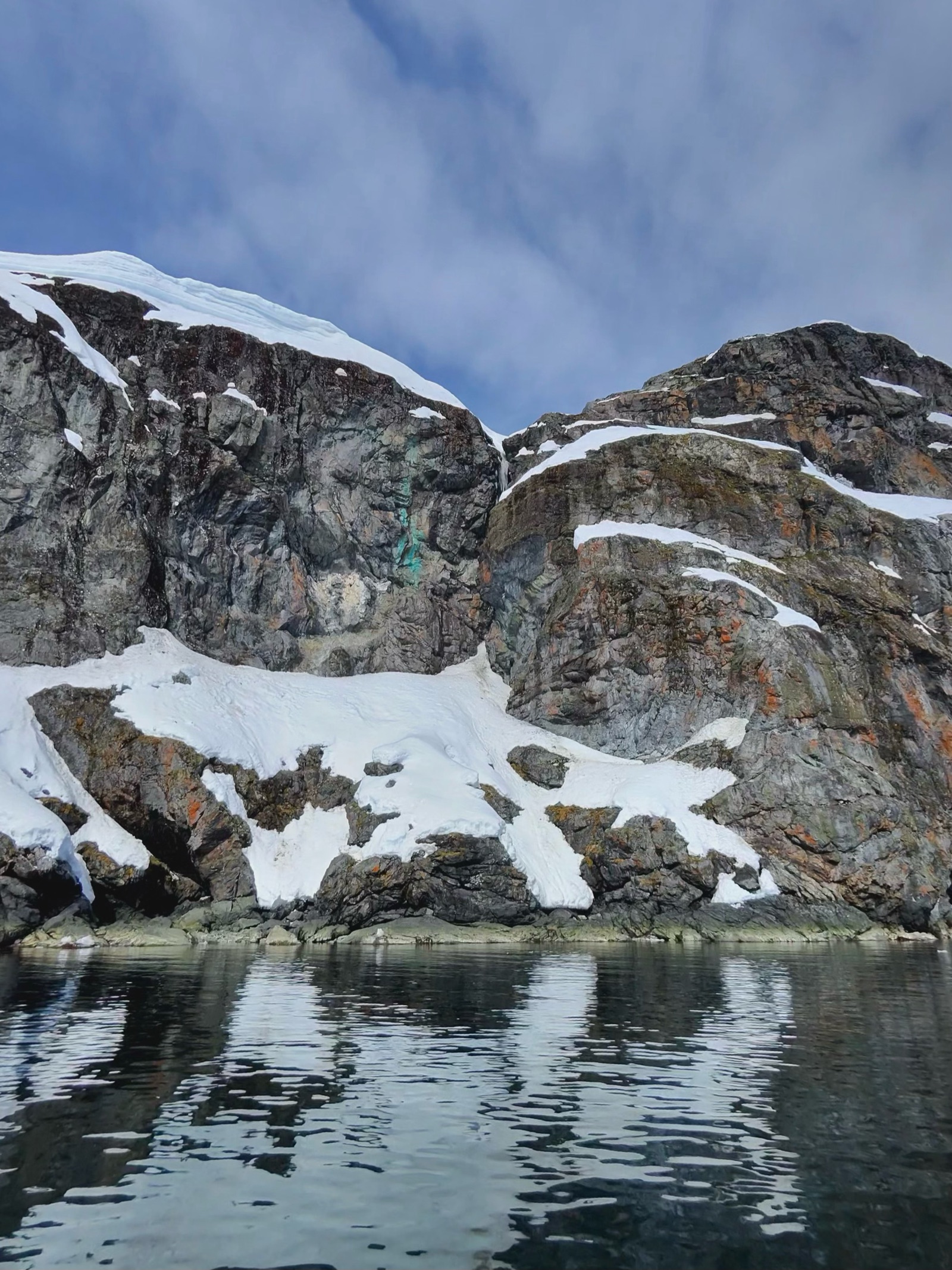 Snow-patched rock cliffs above still water, faces coloured by orange crustose lichen.